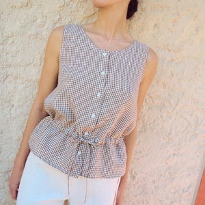 May include: A sleeveless, brown and white gingham top with a button-up front and drawstring waist is paired with white trousers. The top has a scoop neckline and the trousers have a drawstring closure.