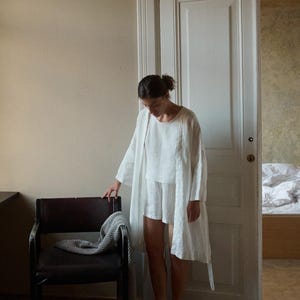 May include: A person wearing a white linen outfit, including a long robe, top, and shorts. The person is standing near a dark brown chair with a gray blanket draped over it. The setting is a room with a white door and beige walls.