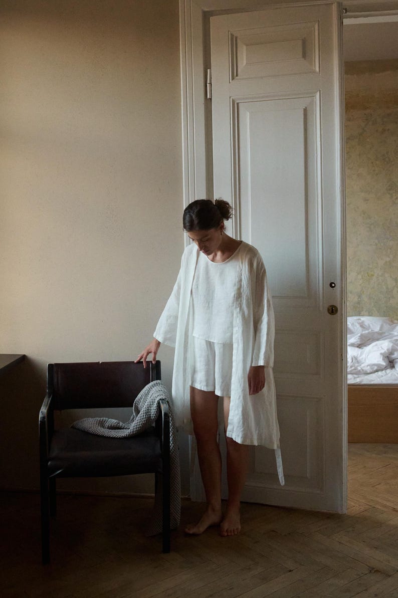 May include: A person wearing a white linen outfit, including a long robe, top, and shorts. The person is standing near a dark brown chair with a gray blanket draped over it. The setting is a room with a white door and beige walls.