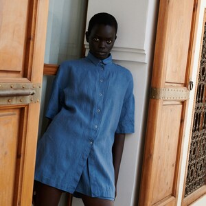 May include: A person wearing a blue linen shirt with short sleeves and a button-down front. The shirt is slightly oversized and has a relaxed fit.