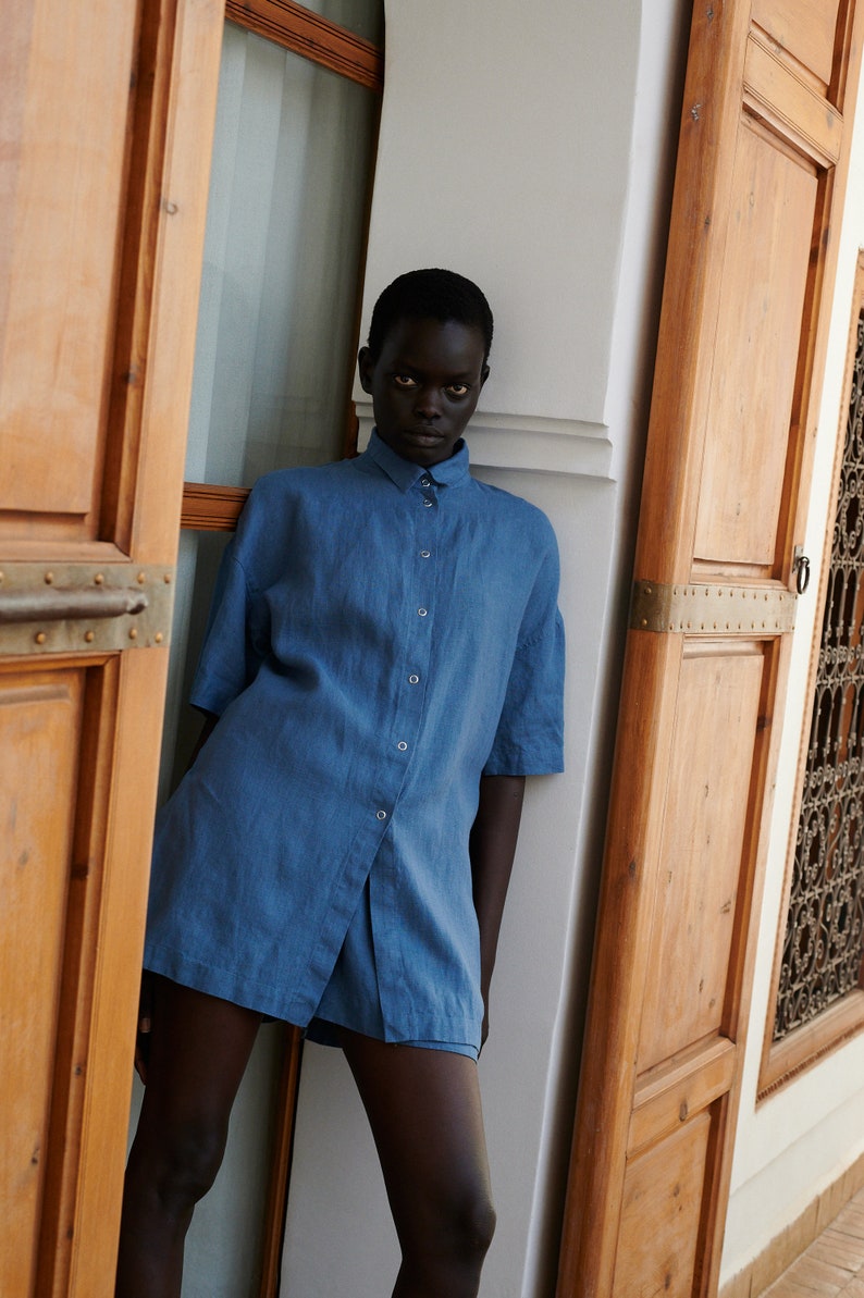 May include: A person wearing a blue linen shirt with short sleeves and a button-down front. The shirt is slightly oversized and has a relaxed fit.