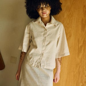 May include: A beige linen shirt and skirt set. The short-sleeved shirt has a button-down front and a collar. The skirt is a midi length with a front slit. The outfit is a neutral color and appears to be made of a textured fabric.