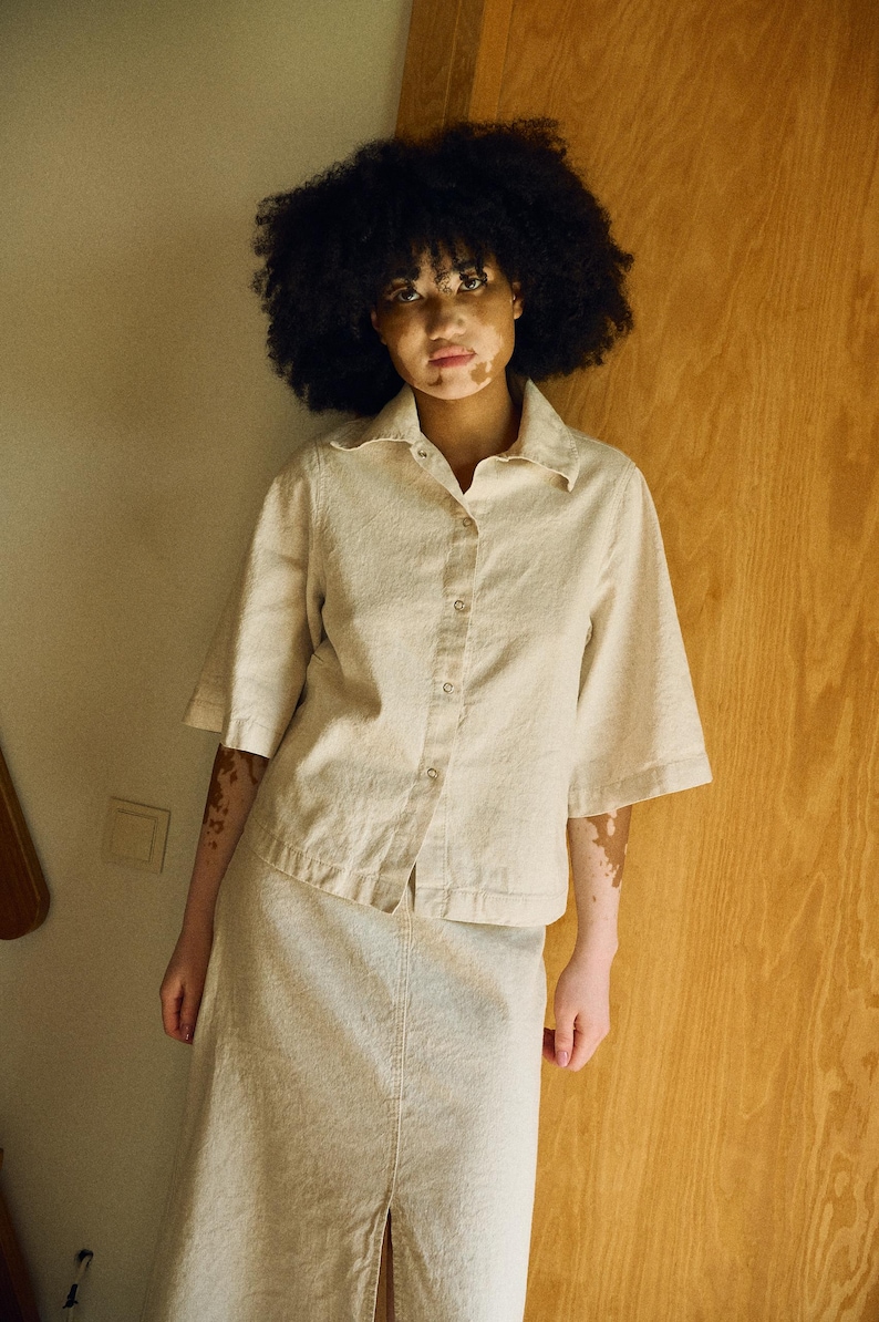 May include: A beige linen shirt and skirt set. The short-sleeved shirt has a button-down front and a collar. The skirt is a midi length with a front slit. The outfit is a neutral color and appears to be made of a textured fabric.