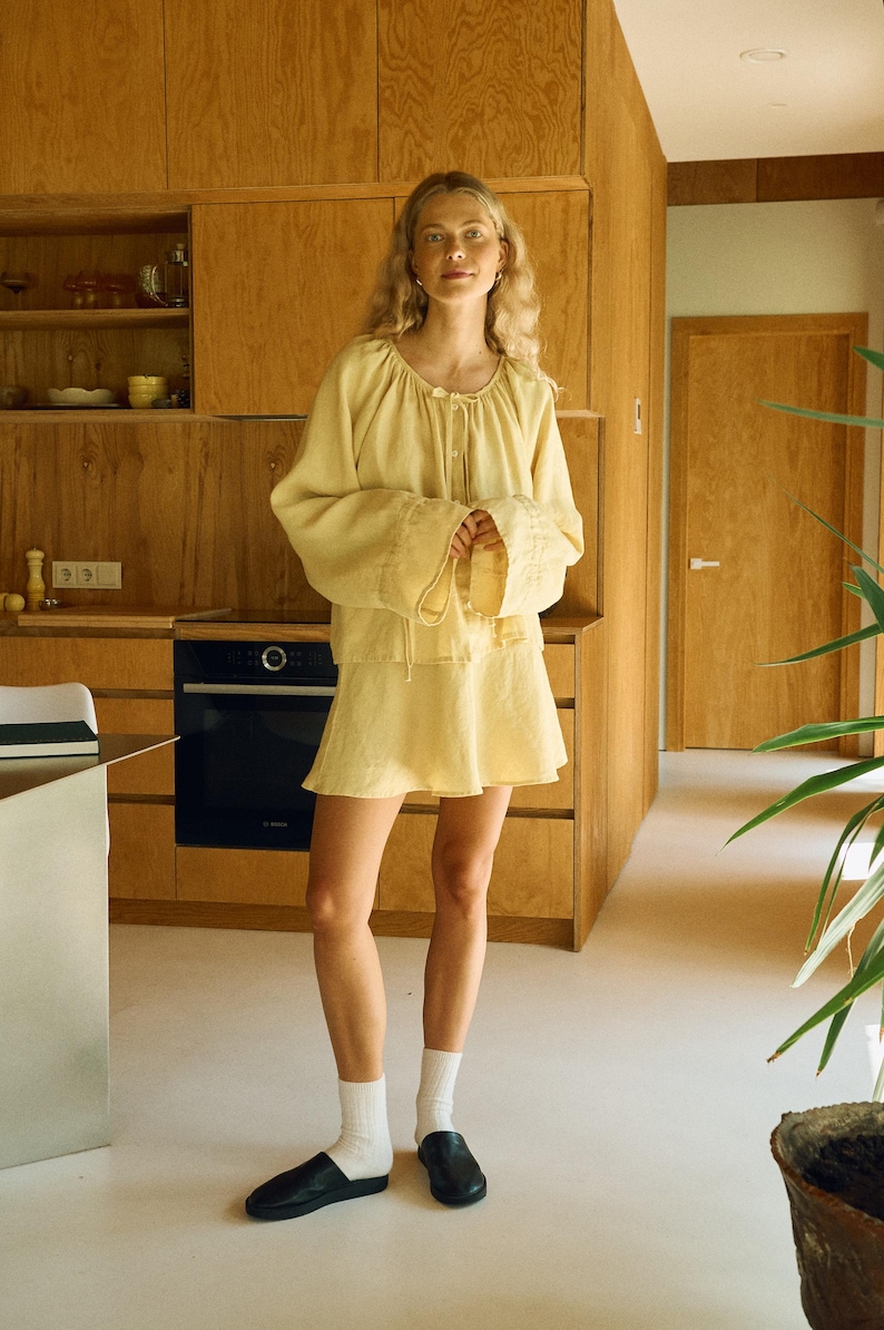 Pode incluir: Uma mulher vestindo um conjunto de linho amarelo claro, composto por uma blusa de manga comprida e uma minissaia. A blusa tem decote redondo e detalhes de cord&atilde;o. Ela est&aacute; usando meias brancas e sapatos pretos. O fundo apresenta arm&aacute;rios de madeira e uma cozinha moderna.