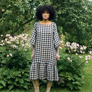 May include: A woman wearing a navy and beige checkered dress with a square neckline and short sleeves. The dress has a ruffled hem. She is standing in a garden with green grass, white flowers, and a large tree in the background.
