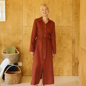 May include: A rust-colored linen dress with long sleeves and a matching belt. The dress has a button-down front and a slit at the hem. The person is wearing black shoes. The background is a light-colored room with wooden paneling.