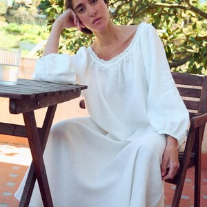 May include: A white linen dress with long sleeves and a lace-trimmed neckline. The dress is worn by a person seated at a wooden table on a brick patio. A glass of water sits on the table. The person is resting their head on their hand.