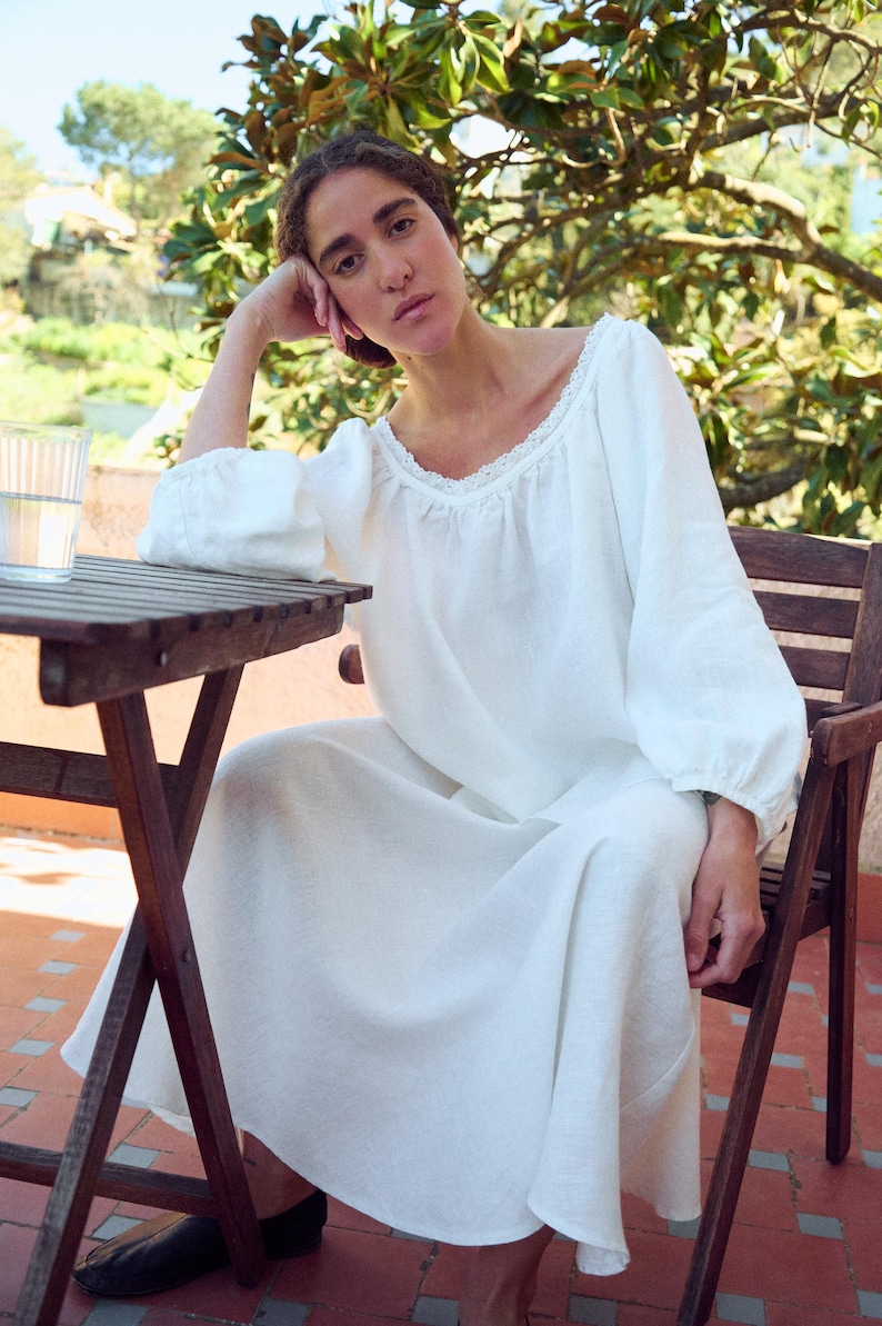 May include: A white linen dress with long sleeves and a lace-trimmed neckline. The dress is worn by a person seated at a wooden table on a brick patio. A glass of water sits on the table. The person is resting their head on their hand.