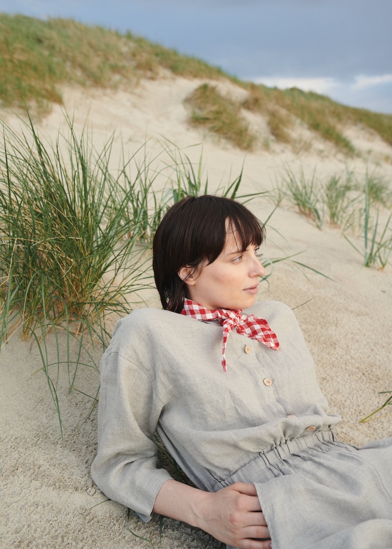 May include: A woman wearing a light gray linen dress with a red and white gingham scarf. She is lying on a sandy beach with green grass in the background.