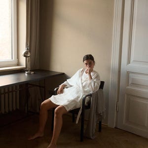 May include: A white linen robe with a belt, worn while seated in a black chair. A patterned blanket is draped over the chair. The room has a desk with a lamp and a window with a curtain.