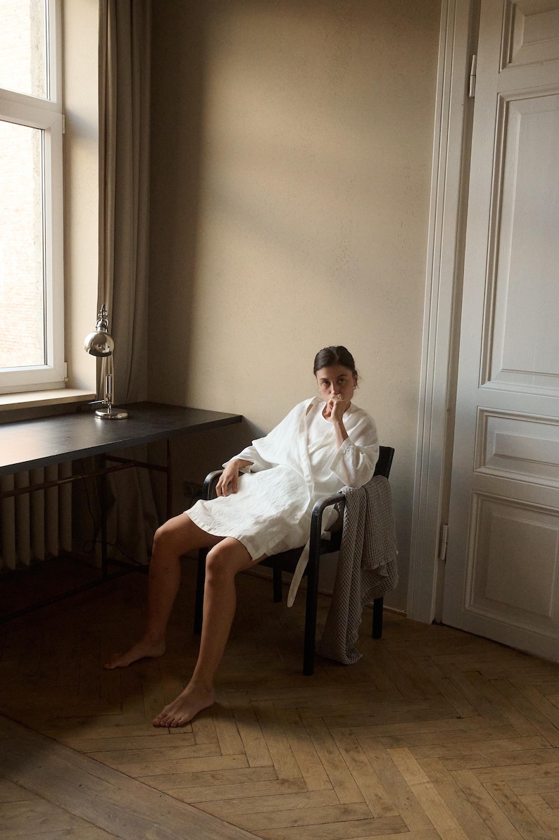 May include: A white linen robe with a belt, worn while seated in a black chair. A patterned blanket is draped over the chair. The room has a desk with a lamp and a window with a curtain.