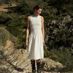 May include: A white sleeveless dress with a smocked bodice and a midi length. The dress is worn by a person standing on a rocky path in a natural setting.