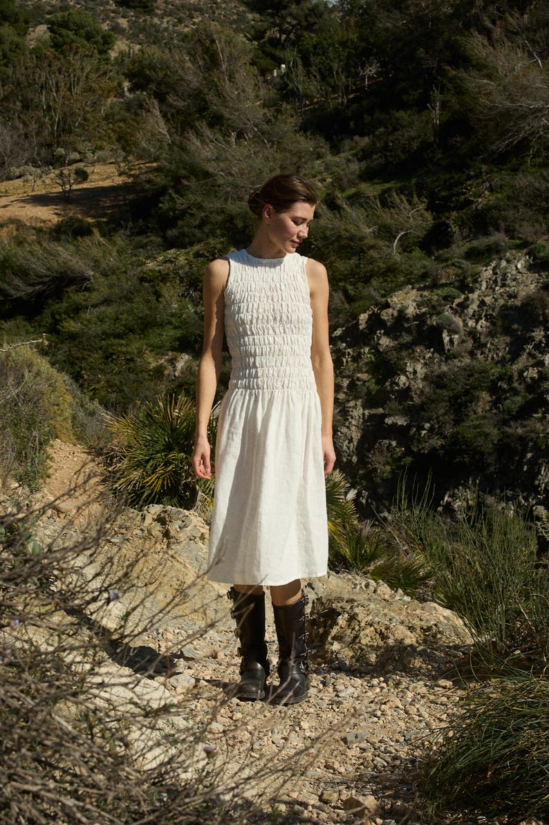 May include: A white sleeveless dress with a smocked bodice and a midi length. The dress is worn by a person standing on a rocky path in a natural setting.
