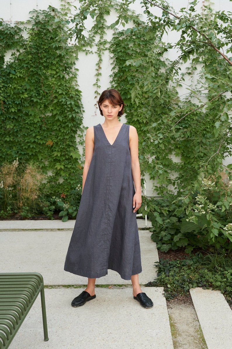 Puede incluir: Una mujer con un vestido largo sin mangas de color gris oscuro con escote en V. Est&aacute; de pie en un camino de piedra con plantas verdes en el fondo.