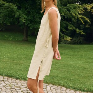 May include: A sleeveless, light beige linen dress with a square neckline and side slits. The dress features tie closures on the side and is paired with a matching headscarf. The model is wearing black leather slides.
