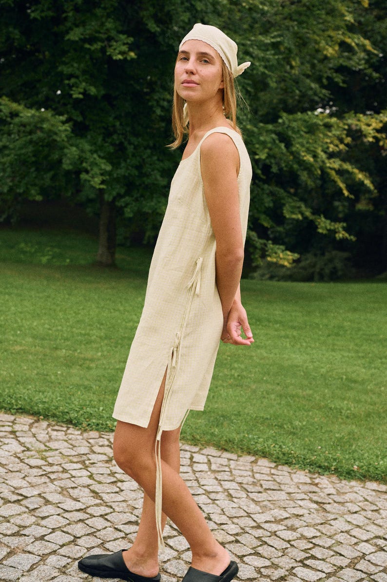 May include: A sleeveless, light beige linen dress with a square neckline and side slits. The dress features tie closures on the side and is paired with a matching headscarf. The model is wearing black leather slides.