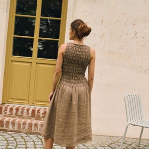 May include: A sleeveless, brown dress with a textured bodice and a flowing skirt. The dress is knee-length and appears to be made of linen or a similar natural fabric. The model is wearing black flats. The background includes a yellow door and a white wall.