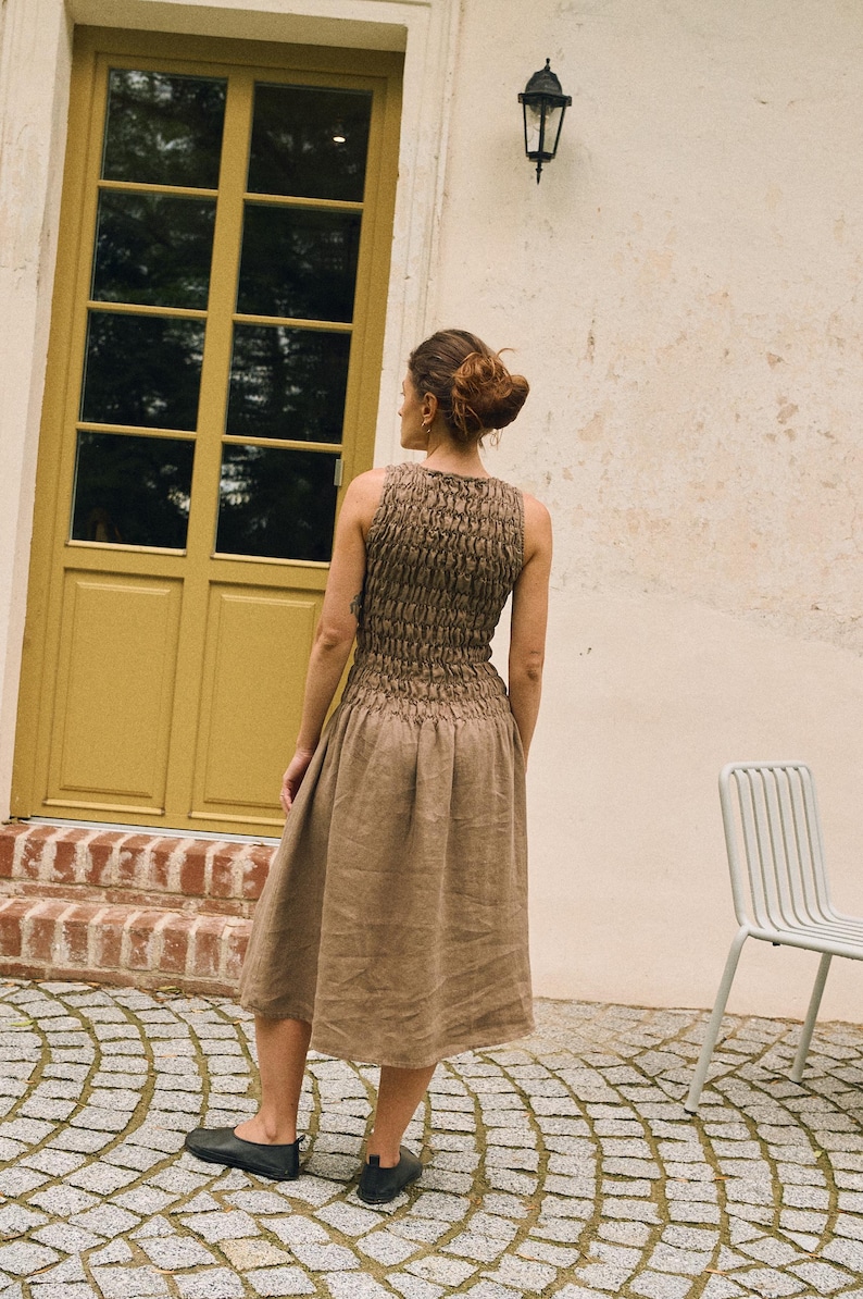 May include: A sleeveless, brown dress with a textured bodice and a flowing skirt. The dress is knee-length and appears to be made of linen or a similar natural fabric. The model is wearing black flats. The background includes a yellow door and a white wall.