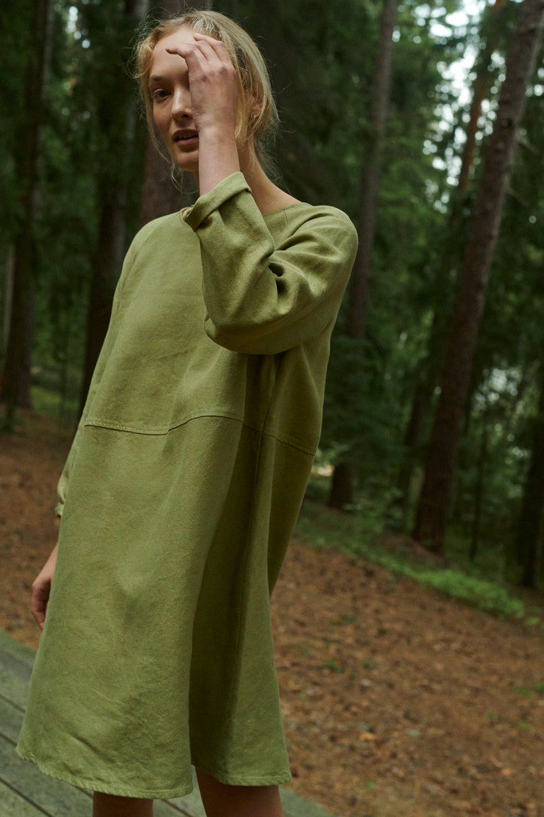 Peut inclure: Une femme portant une robe en lin ample de couleur vert olive &agrave; manches longues. Elle se tient dans un environnement forestier.