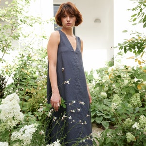 Puede incluir: Una mujer con un vestido de lino sin mangas de color gris oscuro de pie en un jard&iacute;n con flores blancas.