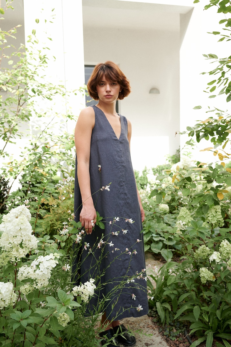Puede incluir: Una mujer con un vestido de lino sin mangas de color gris oscuro de pie en un jard&iacute;n con flores blancas.