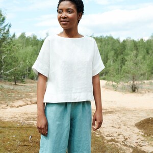 May include: A person wearing a white linen top with short sleeves and light blue linen shorts. The person is standing in a grassy area with trees in the background.