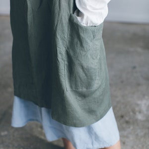 May include: A green linen apron with a large pocket on the front. The apron is worn over a light blue skirt.