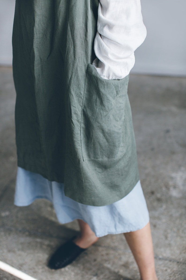 May include: A green linen apron with a large pocket on the front. The apron is worn over a light blue skirt.