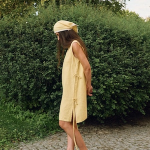 May include: A sleeveless, pale yellow dress with side ties and a matching headscarf. The dress has a simple, relaxed fit and is paired with black flats. The outfit is set against a backdrop of green foliage and a stone path.