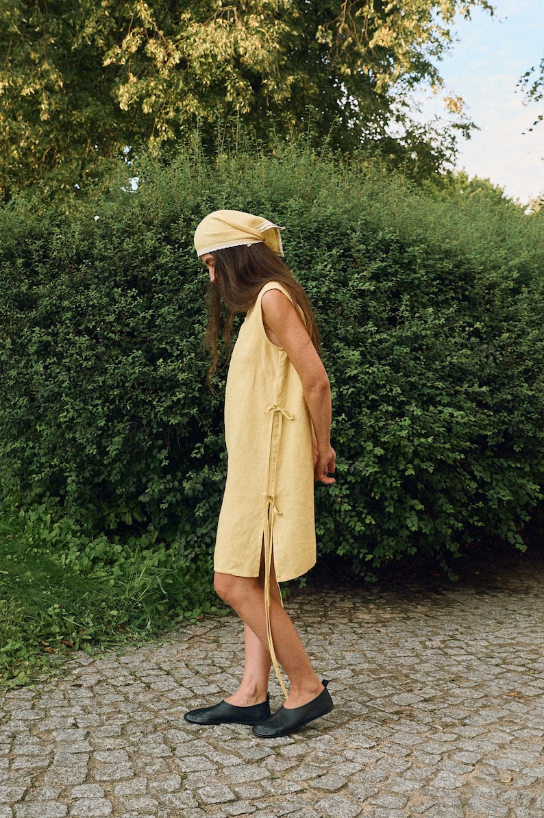 May include: A sleeveless, pale yellow dress with side ties and a matching headscarf. The dress has a simple, relaxed fit and is paired with black flats. The outfit is set against a backdrop of green foliage and a stone path.
