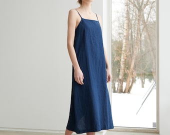 Puede incluir: Una mujer lleva un vestido midi de lino azul oscuro con tirantes finos. Está de pie frente a una ventana con un paisaje nevado en el fondo. Lleva sandalias de cuero negro con hebillas.