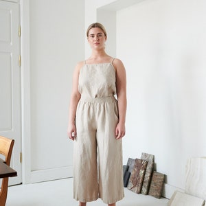 May include: A woman wearing a light beige linen jumpsuit with a wide leg silhouette. The jumpsuit has a square neckline and thin straps. The woman is standing in a white room with a wooden door and a table in the background.