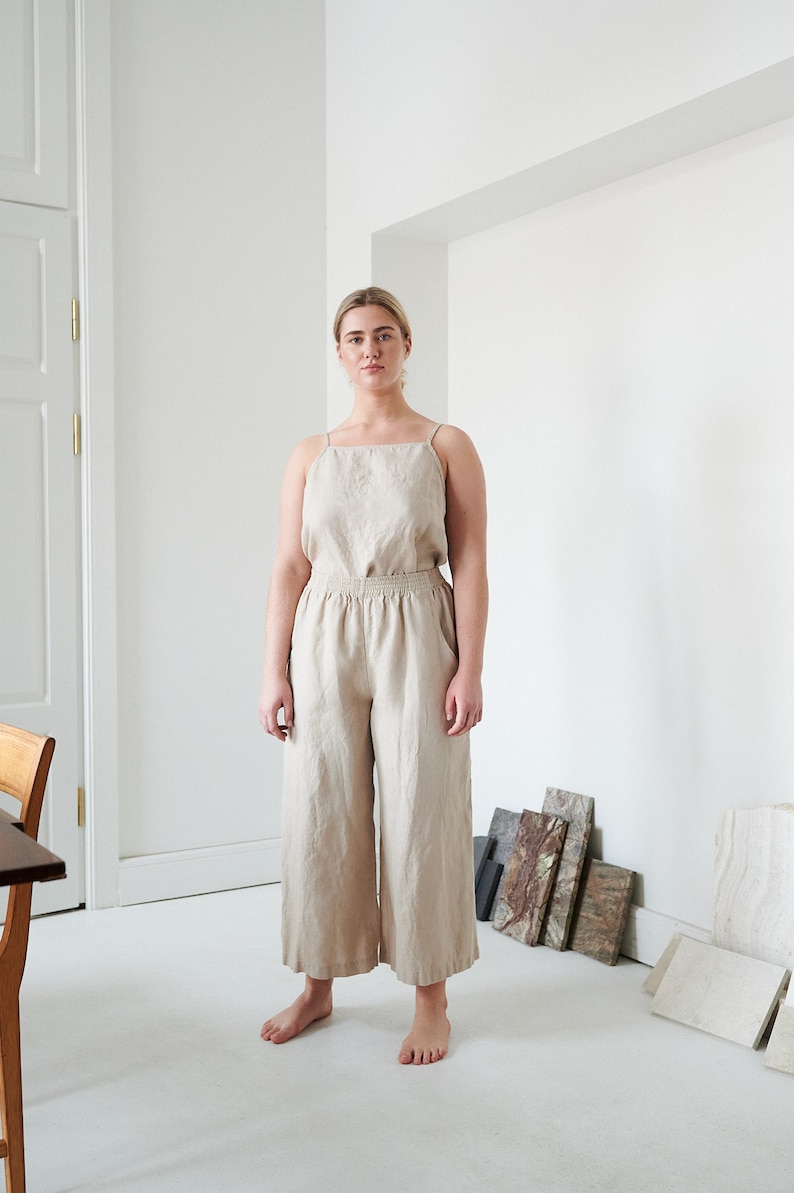May include: A woman wearing a light beige linen jumpsuit with a wide leg silhouette. The jumpsuit has a square neckline and thin straps. The woman is standing in a white room with a wooden door and a table in the background.