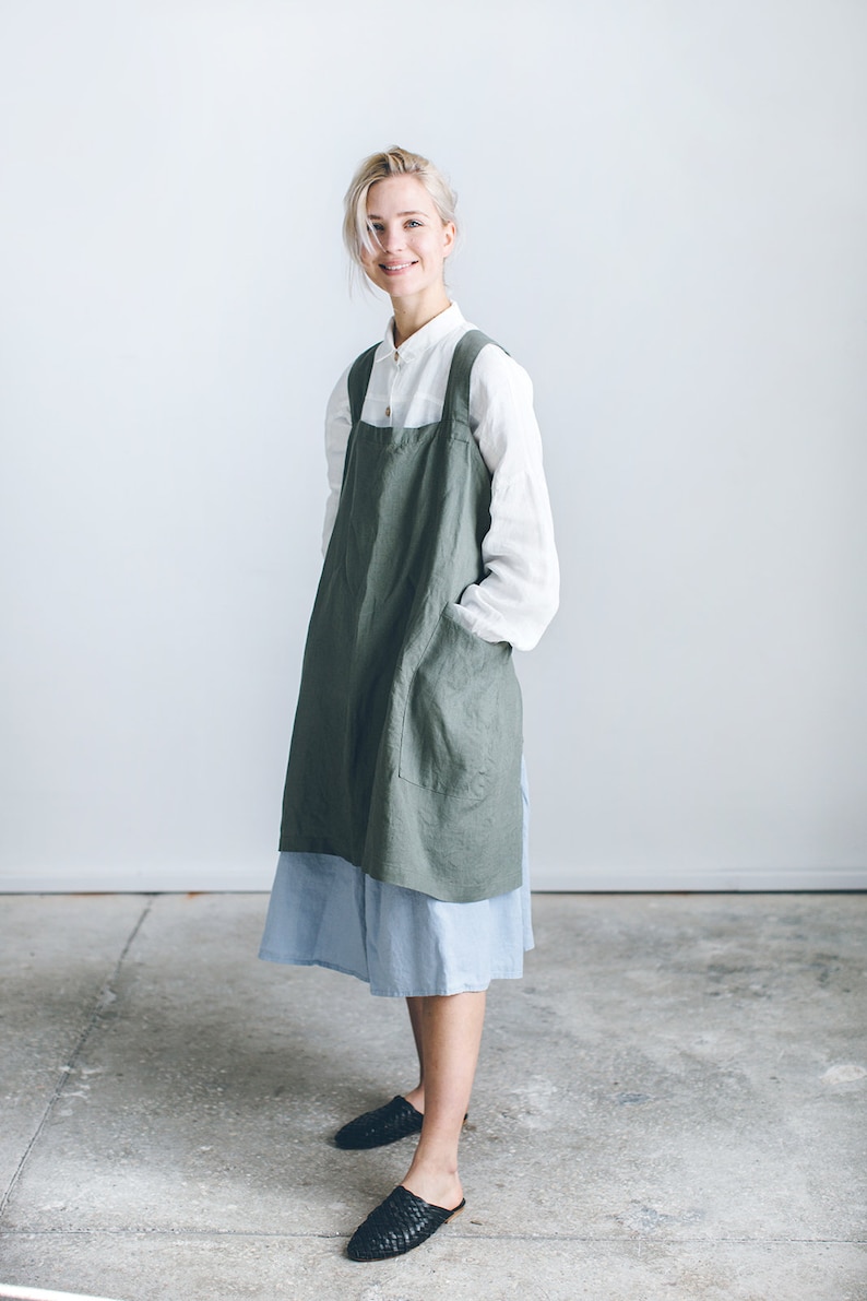 May include: A woman wearing a green linen apron over a white shirt and a light blue skirt. She is standing on a concrete floor and smiling at the camera.