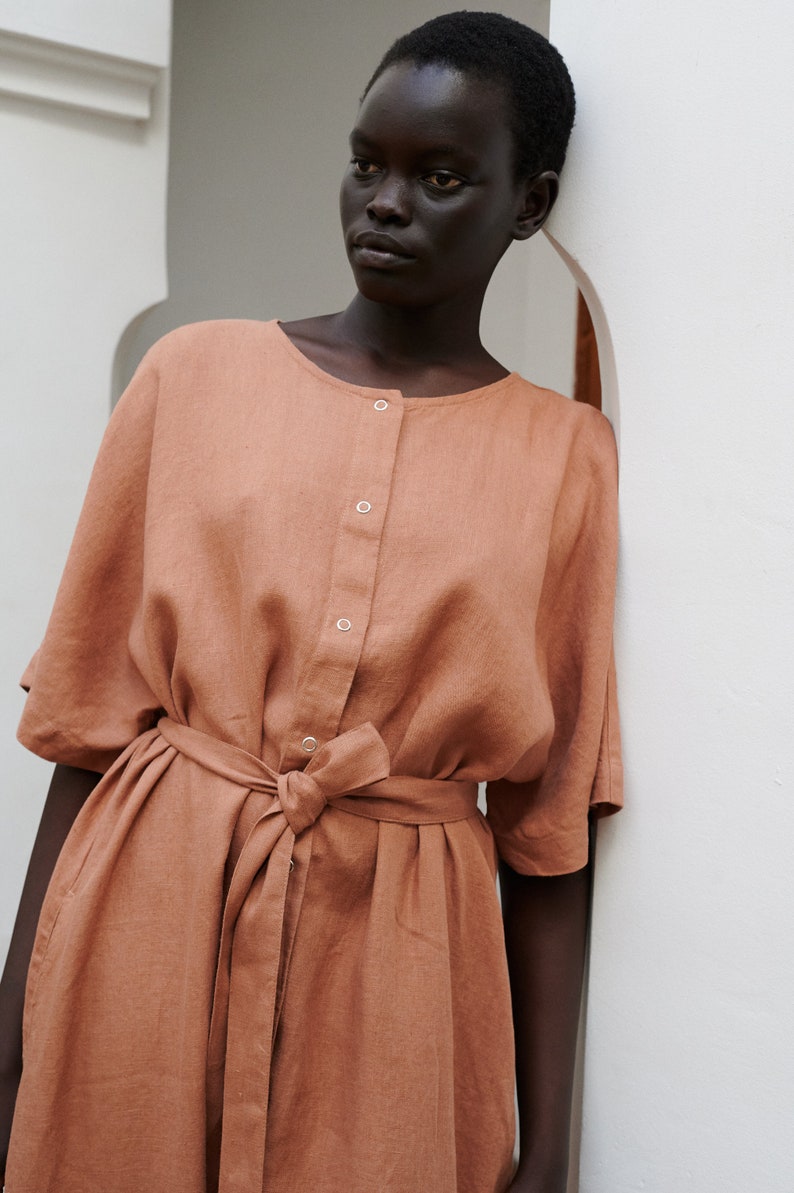 Puede incluir: Un vestido de lino terracota con botones en la parte delantera y un cintur&oacute;n de anudar. El vestido tiene un corte holgado y una falda ligeramente acampanada.