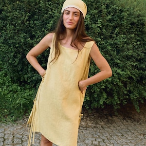 May include: A woman wearing a yellow linen dress and matching headscarf. The sleeveless dress has a square neckline and tie details on the sides. The head covering has a lace trim. The background is a green hedge and a stone path.