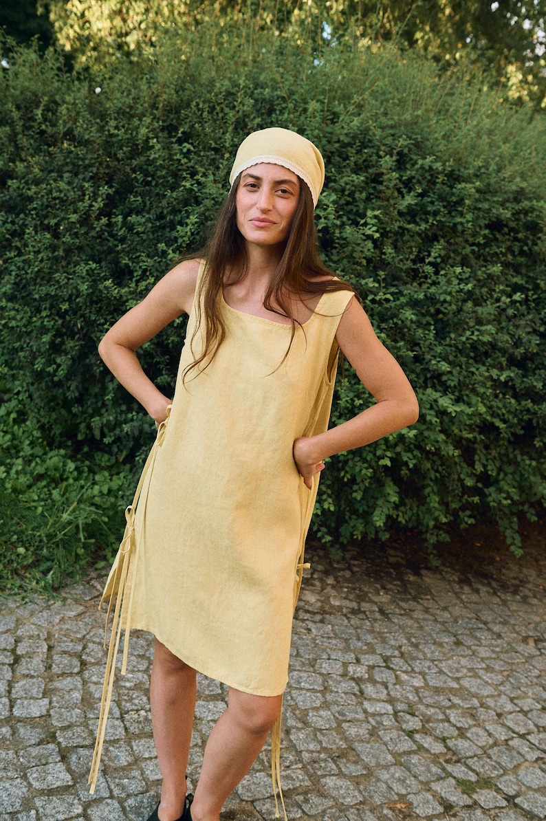 May include: A woman wearing a yellow linen dress and matching headscarf. The sleeveless dress has a square neckline and tie details on the sides. The head covering has a lace trim. The background is a green hedge and a stone path.