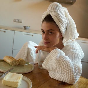 以下が含まれることがあります： 白いワッフル生地のローブと頭に巻いた白いタオルをつけた女性が木製のテーブルに座っています。彼女はカメラを見ています。テーブルにはパンとバターの皿があります。