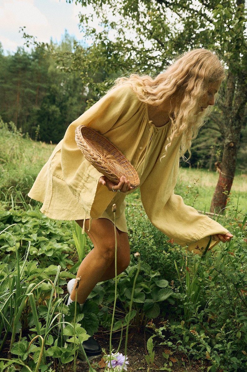 Pode incluir: Uma pessoa vestindo um vestido de linho amarelo e segurando uma cesta de vime em um jardim. A pessoa est&aacute; colhendo plantas em meio &agrave; folhagem verde. O vestido tem mangas compridas e decote com cord&atilde;o. A cena &eacute; ao ar livre com &aacute;rvores ao fundo.