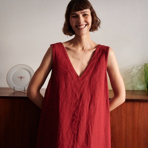 May include: A woman wearing a sleeveless, deep red linen dress with a V-neckline. She is smiling and standing in front of a wooden cabinet.