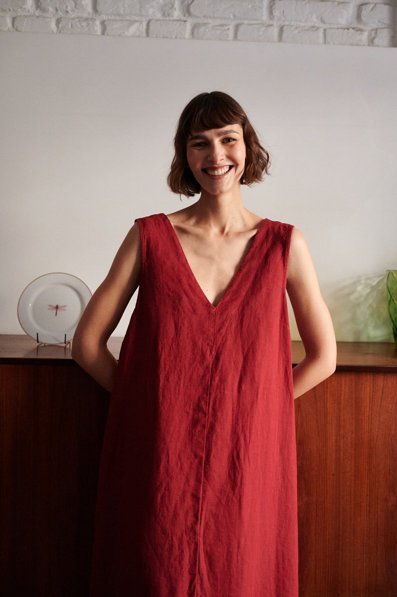 May include: A woman wearing a sleeveless, deep red linen dress with a V-neckline. She is smiling and standing in front of a wooden cabinet.