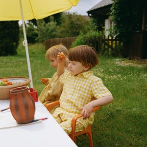 Könnte beinhalten: Zwei Kinder sitzen unter einem gelben Sonnenschirm an einem Tisch in einem grünen Garten. Ein Kind trägt ein gelbes Vichy-Outfit und sitzt auf einem orangefarbenen Stuhl. Ein Keramiktopf und Buntstifte stehen auf dem Tisch.