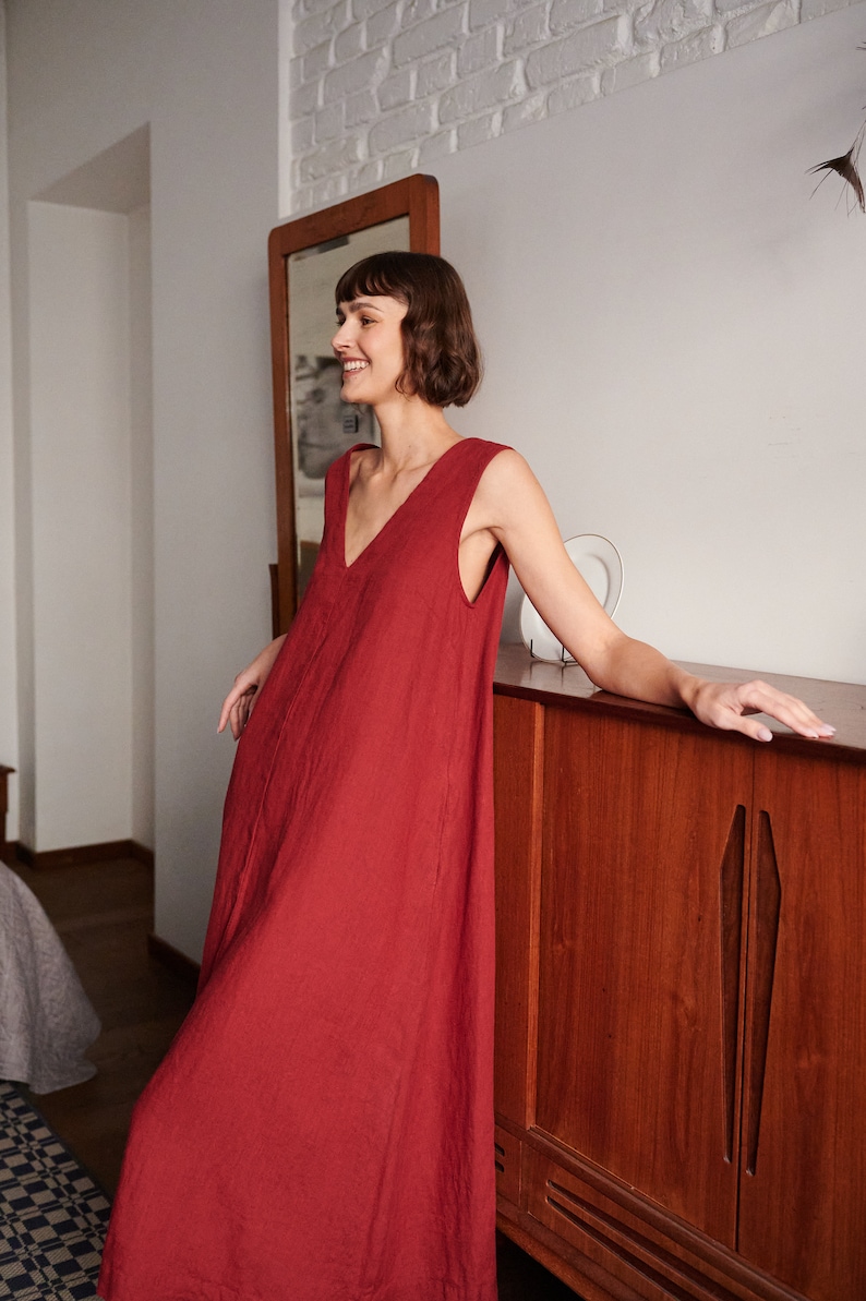 May include: A woman wearing a sleeveless, red linen dress with a V-neckline. She is standing next to a wooden cabinet with a closed door.