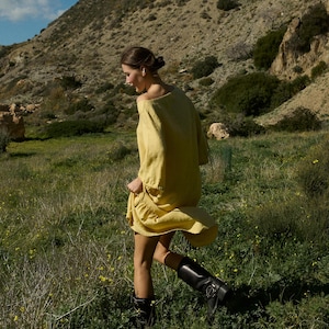 May include: A woman wearing a yellow linen dress and black boots walks through a field of green grass and yellow flowers.