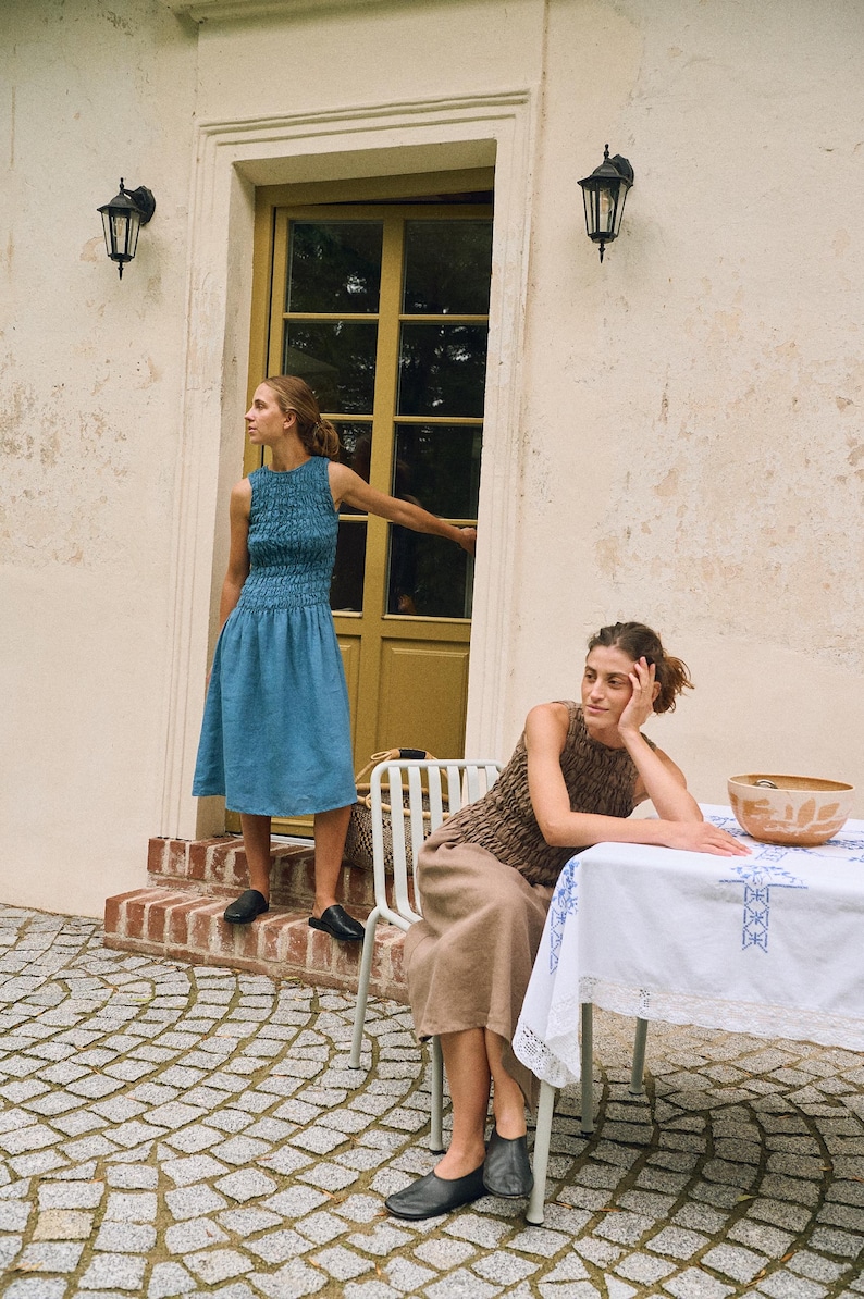 May include: Two women in dresses pose outdoors. One wears a blue sleeveless dress with a textured bodice and a flared skirt. The other wears a brown textured top and a beige skirt. They are near a table with a white tablecloth.