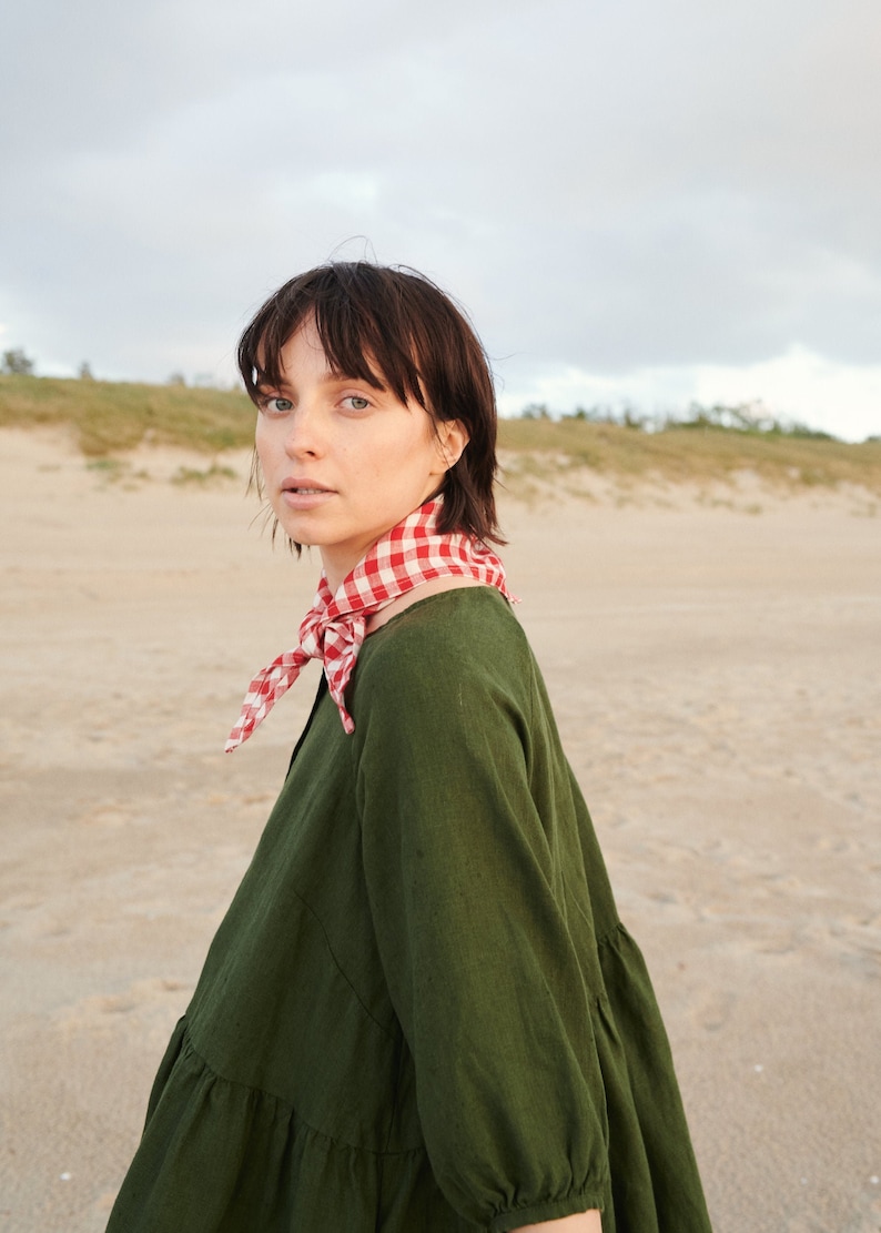 May include: A woman wearing a green dress with a red and white checkered scarf. She is standing on a sandy beach with dunes in the background.