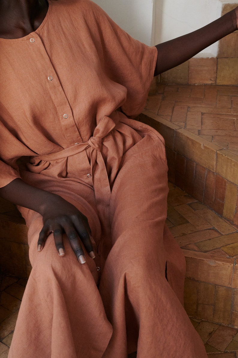 Puede incluir: Un vestido de lino terracota con botones en la parte delantera y un cintur&oacute;n de lazo. El vestido tiene un corte holgado y una falda fluida.