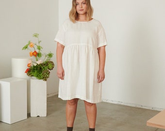 May include: A woman wearing a white linen dress with short sleeves and a gathered skirt. She is standing in a white room with a plant in a white pot.