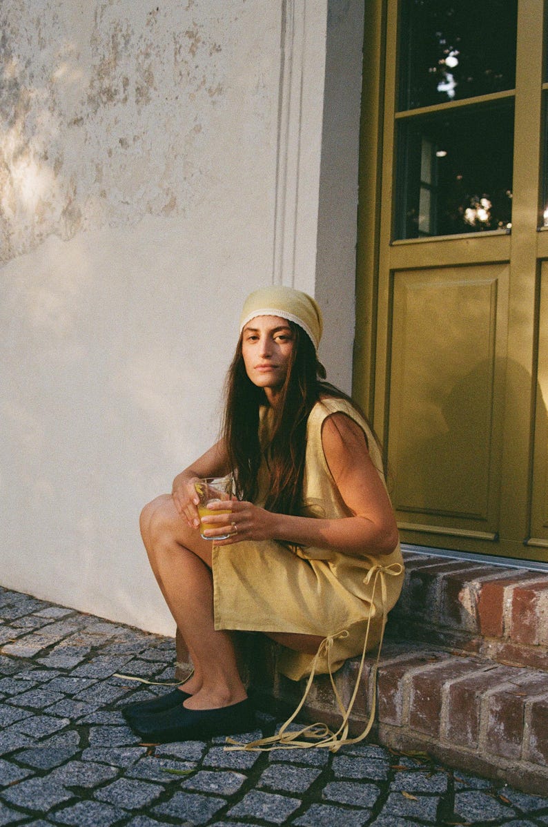 May include: A woman wearing a yellow dress and headscarf, holding a glass of orange liquid. She is sitting on a brick step near a yellow door and a white wall. She is wearing black shoes.