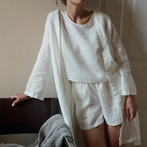 May include: A white linen loungewear set, including a long-sleeved top, shorts, and a long, open-front robe. The outfit is lightweight and appears comfortable. A gray textured blanket is draped over a dark brown chair.